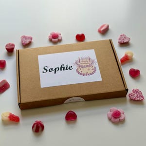 May include: A brown cardboard gift box with a white label that says "Sophie" and a cake illustration. The box is surrounded by various candies, including heart-shaped and flower-shaped gummies in shades of pink and red.