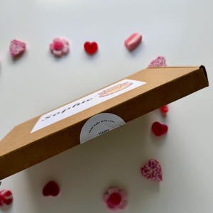 May include: A brown cardboard box with the name "Sophie" printed on a white label. A round sticker with the words "Hand Made With Love Happy" is on the box. Scattered around the box are pink and red candies in various shapes.