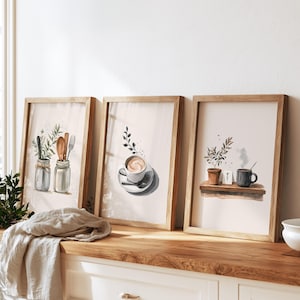 Puede incluir: Tres láminas de acuarela enmarcadas en madera clara. La lámina de la izquierda muestra utensilios de cocina en dos tarros de cristal. La central presenta una taza de café sobre un plato. La de la derecha muestra una pequeña planta en maceta y una taza sobre un estante.