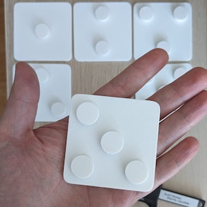 Może przedstawiać: Biały kwadrat z czterema okrągłymi podkładkami samoprzylepnymi. Podkładki są rozmieszczone równomiernie i mają średnicę około 2,5 cm.