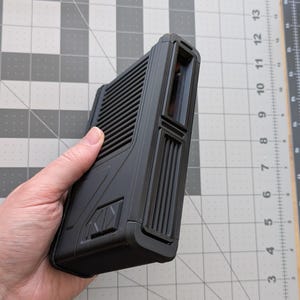 May include: A black, rectangular electronic device with a ridged surface and a small window. The device has a textured design and is held in a hand.