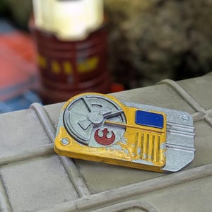 May include: A rectangular, metallic prop with a yellow and silver finish, featuring a red symbol and blue accent. The prop is set on a light gray surface, with a blurred background including a brown cylindrical object.