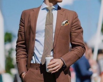 May include: A brown suit with a light blue shirt and patterned tie. The suit has a pocket square and is worn with sunglasses. The person is holding a white can. The background includes other people and a blue sky.