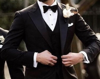 May include: A black tuxedo with a satin lapel, bow tie, and vest. The suit features a white dress shirt and a small floral boutonniere. The person is wearing sunglasses and a wristwatch. The overall look is formal and elegant.
