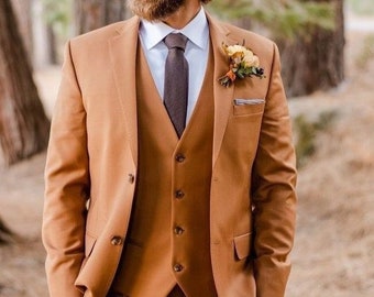 May include: A three-piece suit in a warm, earthy tone. The suit includes a blazer, vest, and trousers. A white dress shirt and brown tie are visible. A floral boutonniere adds a decorative touch. The suit is tailored and well-fitted.