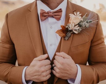 May include: A brown suit with a matching vest and bow tie. The suit has a white dress shirt underneath. A floral boutonniere is pinned to the lapel. The suit is a solid color and appears to be made of a textured fabric.