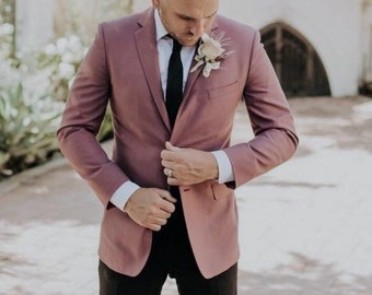 Puede incluir: Chaqueta de traje rosa polvoriento, corbata negra y pantalón negro. La chaqueta se lleva sobre una camisa blanca, con una flor prendida en la solapa. La persona se está ajustando la chaqueta.