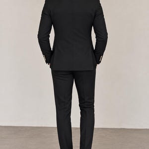 May include: A solid black suit consisting of a tailored blazer with gold-toned buttons on the cuffs and matching slim-fit trousers. The blazer features a structured back panel and a white shirt collar visible at the neckline, paired with black shoes.