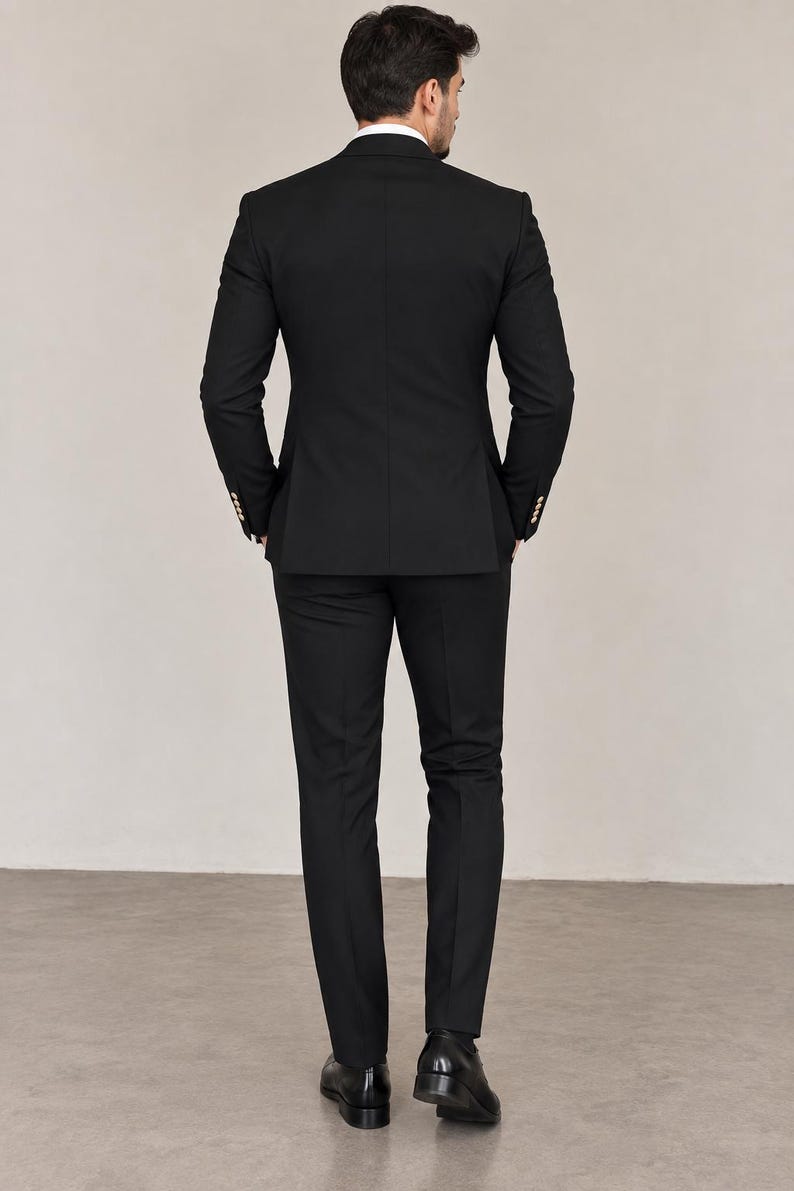 May include: A solid black suit consisting of a tailored blazer with gold-toned buttons on the cuffs and matching slim-fit trousers. The blazer features a structured back panel and a white shirt collar visible at the neckline, paired with black shoes.