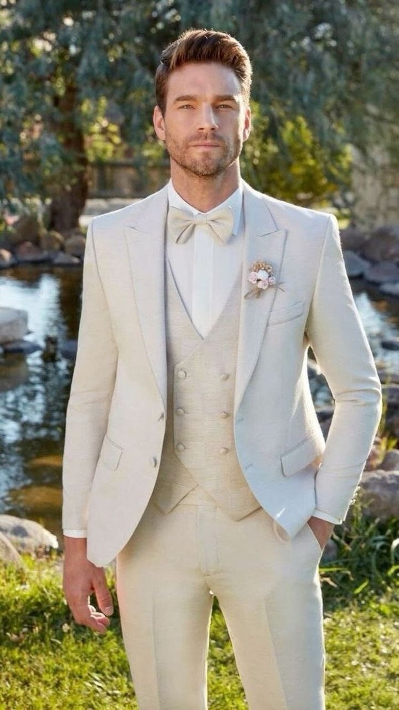 May include: A three-piece suit in a light beige color. The suit includes a jacket, vest, and trousers. A white dress shirt and a beige bow tie complete the look. A small floral boutonniere is pinned to the lapel.
