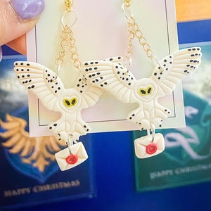 May include: A pair of gold chain earrings with white owl charms holding red envelopes. The owls have black eyes and black spots on their wings. The earrings are on a white card with the text "Forever Green Jewellery".