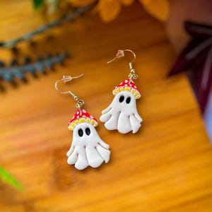Può includere: Un paio di orecchini a fantasma fatti a mano. Ogni orecchino presenta un fantasma bianco con occhi neri e un cappello a fungo rosso a pois bianchi. Gli orecchini pendono da ganci color oro.