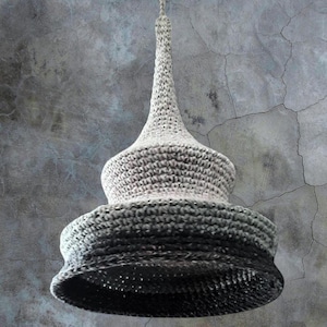 以下が含まれることがあります： 白から黒へのグラデーションで編まれた、クロシェのペンダントライト。ライトは、テクスチャのあるグレーの壁の天井から吊り下げられています。画像の下部には、「CUICI unconventional crochet Torino」というテキストが表示されています。