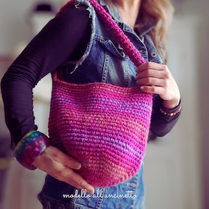 Può includere: Una borsa a mano in crochet rosa e viola con un giubbotto di jeans e un braccialetto arcobaleno. La borsa ha una tracolla lunga ed è realizzata con un punto a trama.