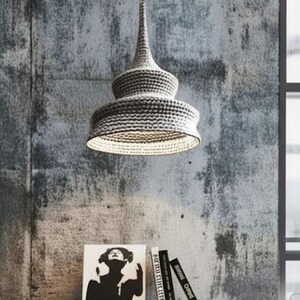 May include: A gray woven pendant light hangs above a white distressed table. The light fixture has a conical top and a flared, tiered shade. Books and a framed print sit on the table. The background is a textured gray wall.