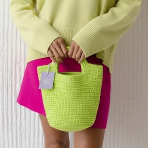 Può includere: Una donna indossa un maglione verde chiaro con scollo a V e una gonna rosa acceso, tenendo in mano una borsa tote all'uncinetto verde lime. La borsa ha un'etichetta attaccata. Lo sfondo è un muro bianco.