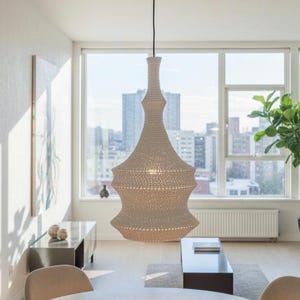 Puede incluir: Una lámpara colgante beige tejida cuelga sobre una mesa redonda de madera en una sala de estar moderna. La lámpara tiene una forma única en capas. La habitación cuenta con grandes ventanales con vistas a la ciudad, una planta en maceta y muebles de tonos neutros.