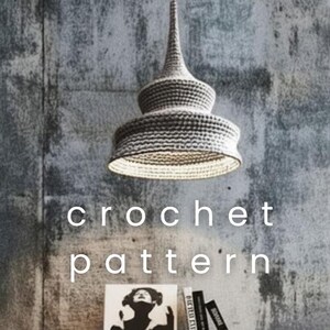 May include: A gray crochet pendant lamp hangs from the ceiling. The lamp has a conical top and a wide, tiered shade. The text "crochet pattern" is displayed below the lamp. A small white table with books and a framed image is below the lamp.