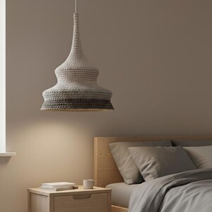 Può includere: Lampada a sospensione con paralume all'uncinetto nei toni del grigio e del bianco. È sospesa sopra un comodino in legno con un cassetto, un libro e una tazza. La camera da letto ha un letto con biancheria da letto grigia.