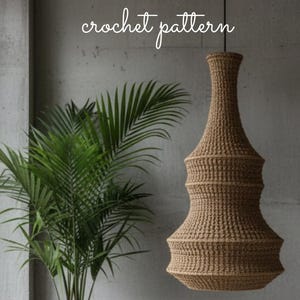 Può includere: Una lampada a sospensione all'uncinetto marrone è appesa davanti a un muro grigio. L'apparecchio ha una forma unica con una base svasata e una parte superiore più stretta. Le parole "crochet pattern" sono scritte sopra la luce.
