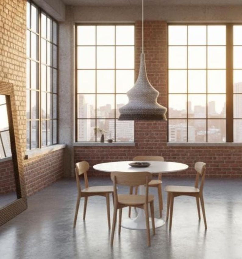May include: A modern dining room with a white round table and four wooden chairs. A woven pendant light hangs above the table. The room has large windows with a city view and exposed brick walls.