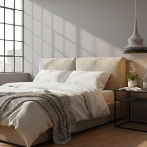 May include: A bedroom scene featuring a bed with a beige headboard, white bedding, and a gray knitted throw. A black side table holds a plant, book, and mug. A gray and black pendant light hangs above. A large window is in the background.