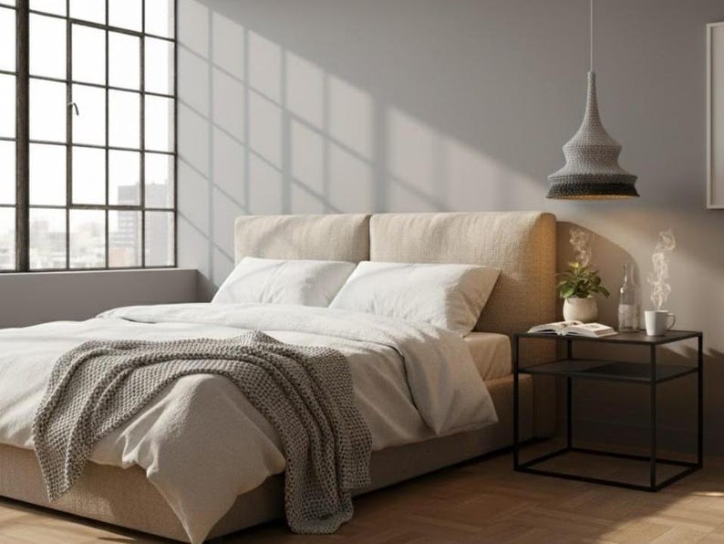 May include: A bedroom scene featuring a bed with a beige headboard, white bedding, and a gray knitted throw. A black side table holds a plant, book, and mug. A gray and black pendant light hangs above. A large window is in the background.