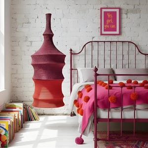 Può includere: Una lampada all'uncinetto rossa e bordeaux è appesa davanti a un muro di mattoni bianchi. Un letto rosa con una coperta con pompon arancioni e una stampa incorniciata con la scritta "Yes girl, you can." è sullo sfondo. Le parole "crochet pattern" sono sopra la lampada.