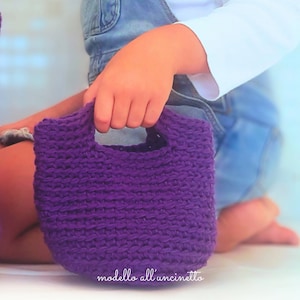 Könnte beinhalten: Eine kleine, handgefertigte, lila Häkeltasche mit Henkel. Die Tasche wird von einer Person gehalten. Der Text "modello all'uncinetto" ist unten rechts im Bild zu sehen.