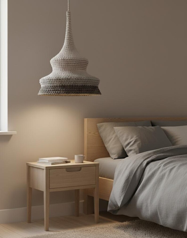 Può includere: Lampada a sospensione con paralume all'uncinetto nei toni del grigio e del bianco. È sospesa sopra un comodino in legno con un cassetto, un libro e una tazza. La camera da letto ha un letto con biancheria da letto grigia.