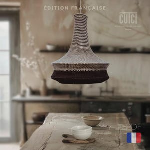 May include: A crocheted lampshade in beige and burgundy with a white label that reads "cuci unconventional crochet Torino" and the text "Édition Française" in the top left corner. The lampshade is hanging from the ceiling in a room with a wooden table and chairs.