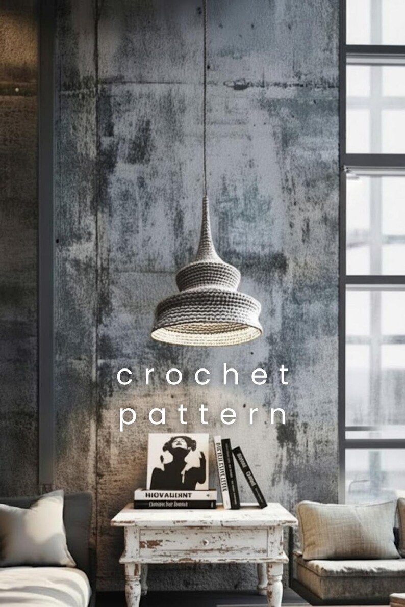 May include: A gray crochet pendant lamp hangs from the ceiling. The lamp has a conical top and a wide, tiered shade. The text "crochet pattern" is displayed below the lamp. A small white table with books and a framed image is below the lamp.