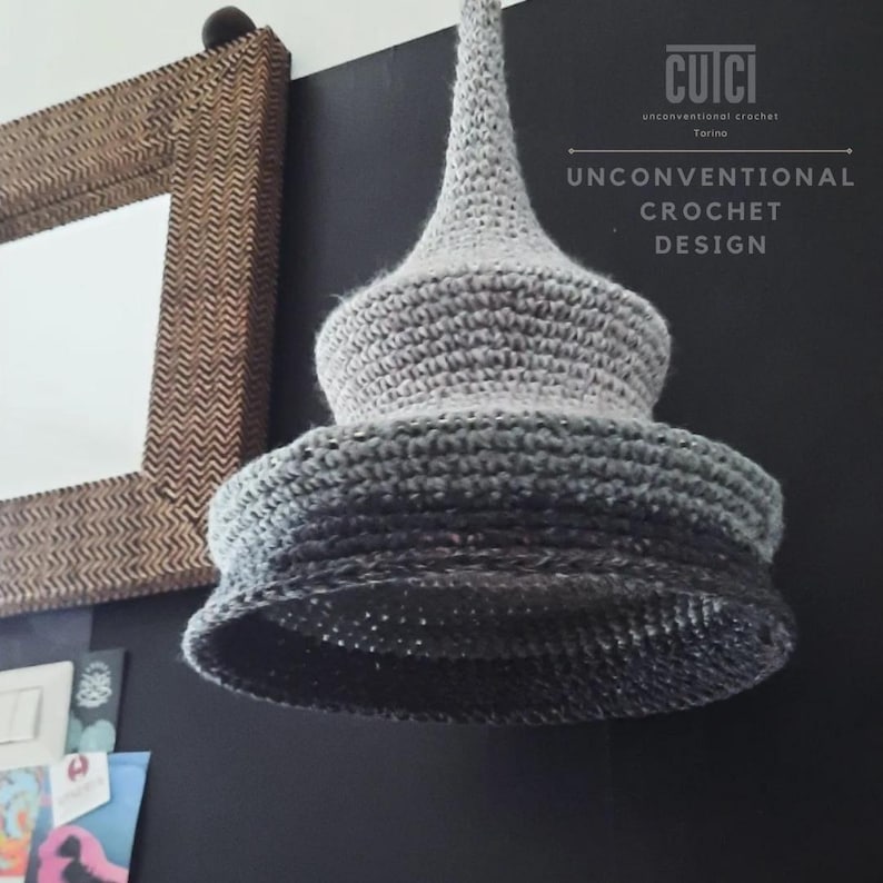 May include: A gray, handmade crochet lampshade with a unique, layered design. The lampshade has a conical top and a flared bottom, with a gradient of gray shades. The text "CUICI UNCONVENTIONAL CROCHET DESIGN" is visible on the right side of the image.