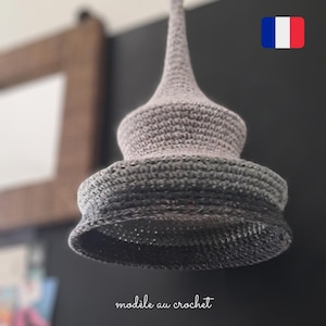 以下が含まれることがあります： 尖った頂上と幅広の丸いベースを持つグレーのクロシェ編みランプシェード。ランプシェードは天井から吊り下げられており、表面にテクスチャがあります。画像の下部に「modèle au crochet」というテキストが表示されています。