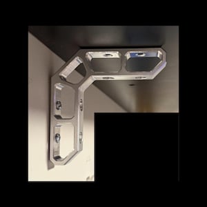 May include: An L-shaped silver metal shelf bracket featuring a structural skeletonized design with multiple rectangular cutouts and pre-drilled circular holes for mounting screws, attached to a flat surface at a ninety-degree angle.