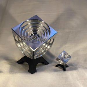 Puede incluir: Dos esculturas geométricas plateadas. La más grande tiene forma de cubo con diseños circulares concéntricos en el interior, montada sobre una base negra. Una versión más pequeña del mismo diseño está al lado.