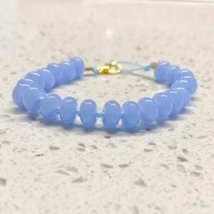 Puede incluir: Un brazalete de cuentas azul claro con un cierre dorado y un cordón azul claro.