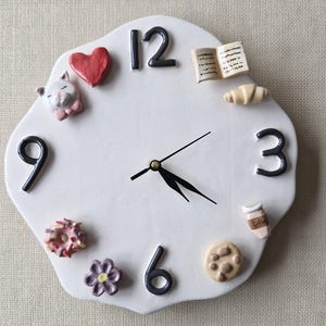 Puede incluir: Reloj de pared de cerámica blanca con números y manecillas negras. Elementos decorativos: corazón rojo, gato, libro, cruasán, donut, flor, galleta y taza de café.