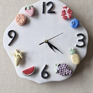 Puede incluir: Reloj de pared blanco con diseño de frutas, números y manecillas negras. La esfera del reloj está adornada con ilustraciones de frutas coloridas, incluyendo fresa, naranja, granada, arándano, piña, uvas, sandía y plátano. El reloj tiene una forma irregular.