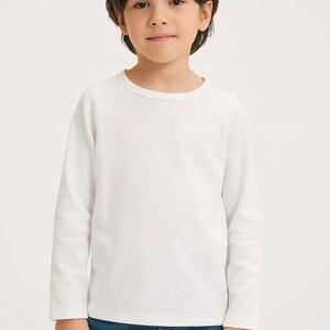 May include: A child wearing a white long-sleeved top and blue jeans. The top has a crew neck and raglan sleeves. The child is standing against a plain background.