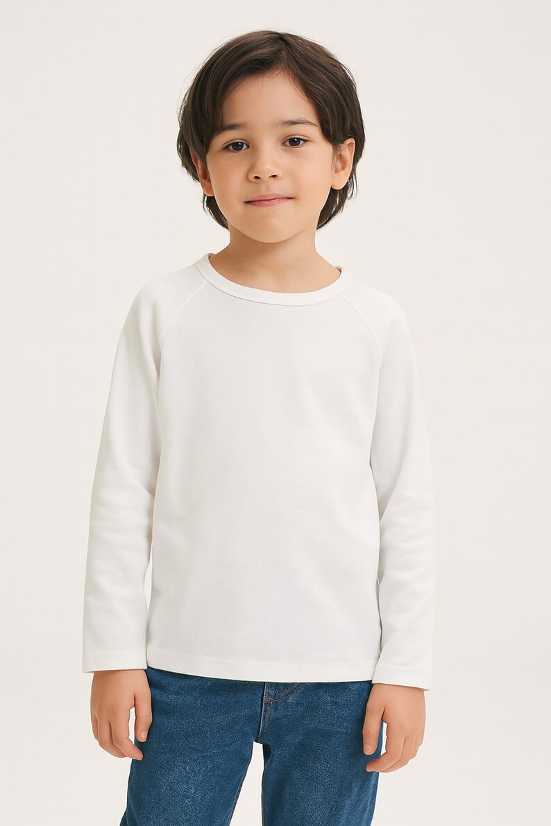 May include: A child wearing a white long-sleeved top and blue jeans. The top has a crew neck and raglan sleeves. The child is standing against a plain background.