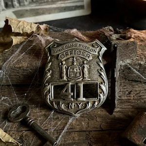 May include: A vintage Sing Sing Prison Officer badge with the number 41, a key, and a padlock. The badge is silver-toned and has the words "Sing Sing Prison Officer" and "NY" on it. A black and white photo of a building is in the background.