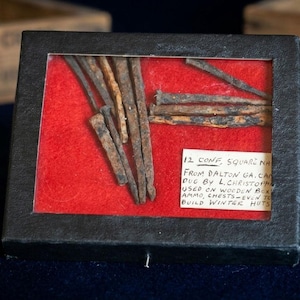 May include: A framed display featuring 12 antique square nails, likely from the Civil War era. The nails are displayed on a red background within a black frame. A handwritten label provides details about their origin and use.