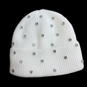 Puede incluir: Un gorro de punto blanco adornado con múltiples pedrerías brillantes. El gorro tiene una textura acanalada y un borde doblado. Las pedrerías están espaciadas uniformemente sobre la superficie del gorro, agregando un toque de glamour al accesorio de invierno.