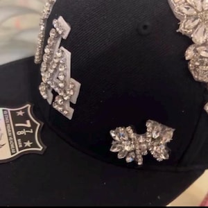 Puede incluir: Una gorra de béisbol negra con un gran adorno de pedrería plateada en la parte delantera. La gorra tiene una visera negra y un parche blanco con el texto "7 1/8 Talla" y "KB EROS" en el lateral.