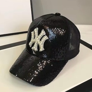 May include: Black baseball cap covered in sequins. The cap features a white and silver emblem on the front. The cap has a curved brim and a mesh back. The cap is a fashion accessory.