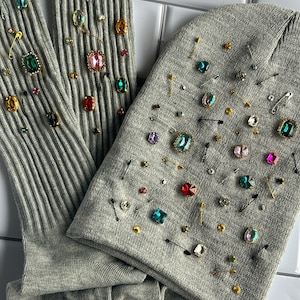 May include: A pair of gray ribbed socks and a gray beanie hat decorated with colorful rhinestones and safety pins.
