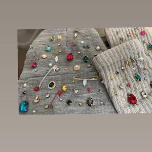 Puede incluir: Dos gorros de punto grises decorados con joyas de colores, pedrería y imperdibles.