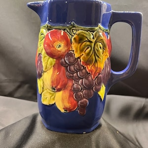 May include: A blue ceramic pitcher with a fruit design. The pitcher has a handle and is decorated with grapes, apples, and pears.
