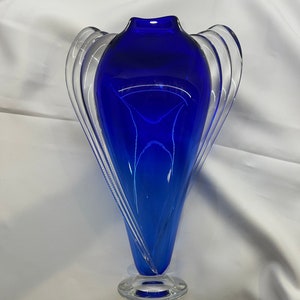 May include: A cobalt blue glass vase with a clear glass base and a unique, flowing design. The vase has a wide mouth and a narrow base.